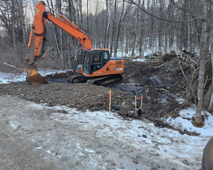 Excavator building a gravel pad beside a new culvert on a wooded site