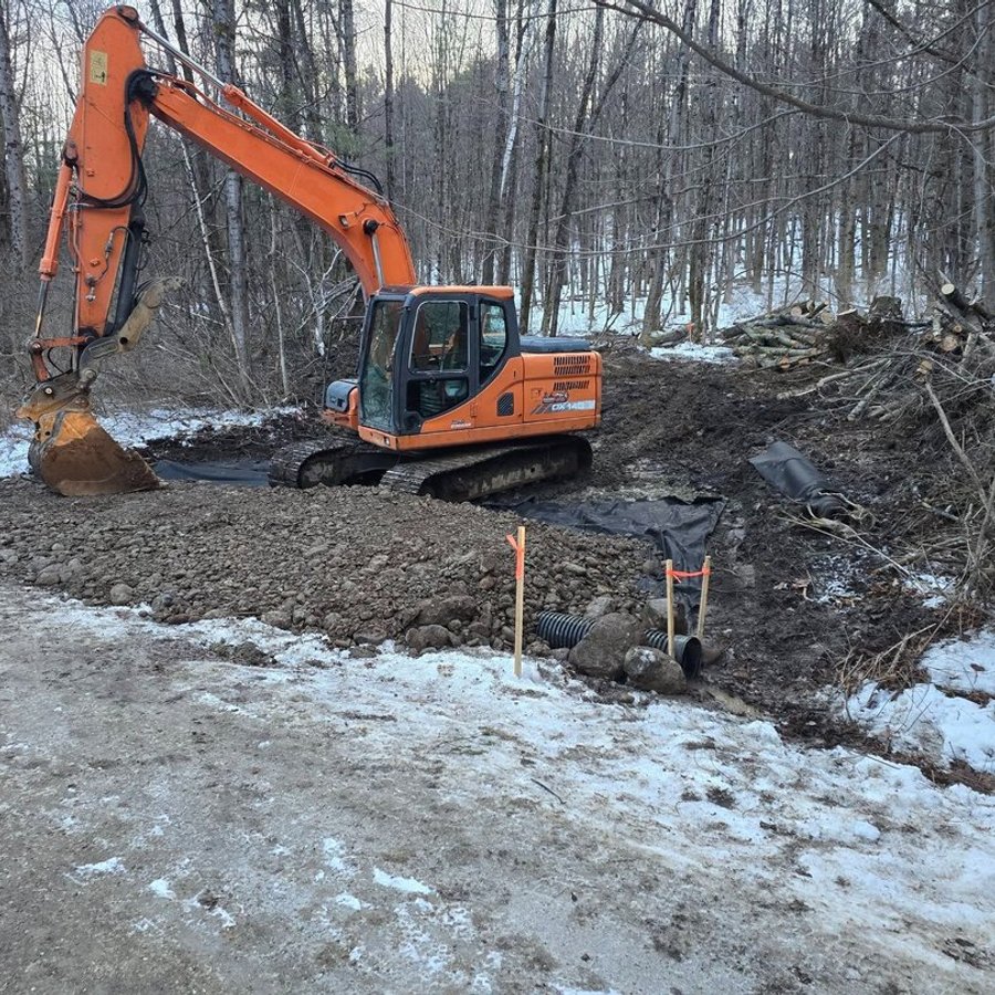 Excavator building a gravel pad beside a new culvert on a wooded site